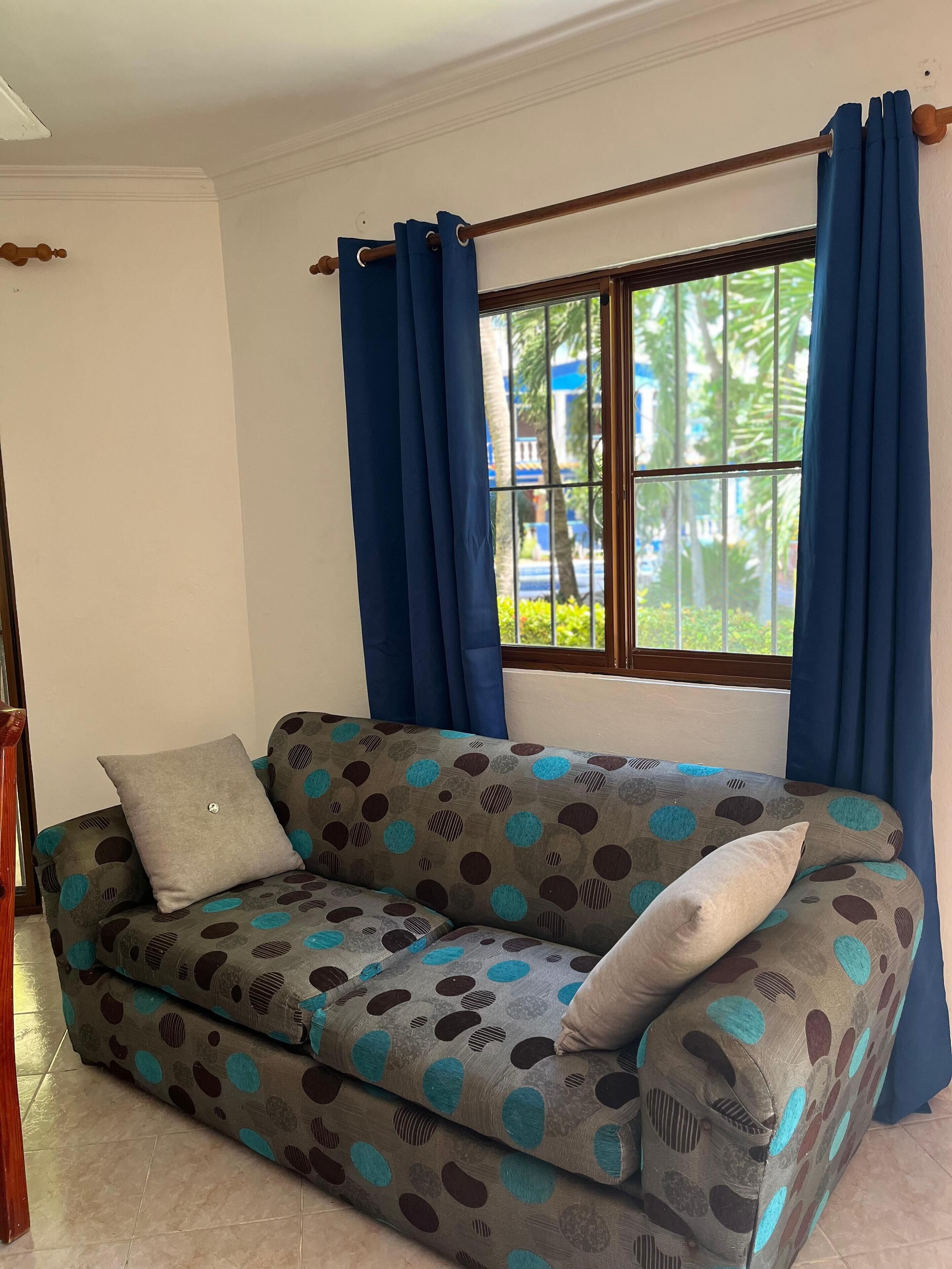 Una sala de estar con un sofá, una ventana con cortinas azules y vistas a zonas verdes.