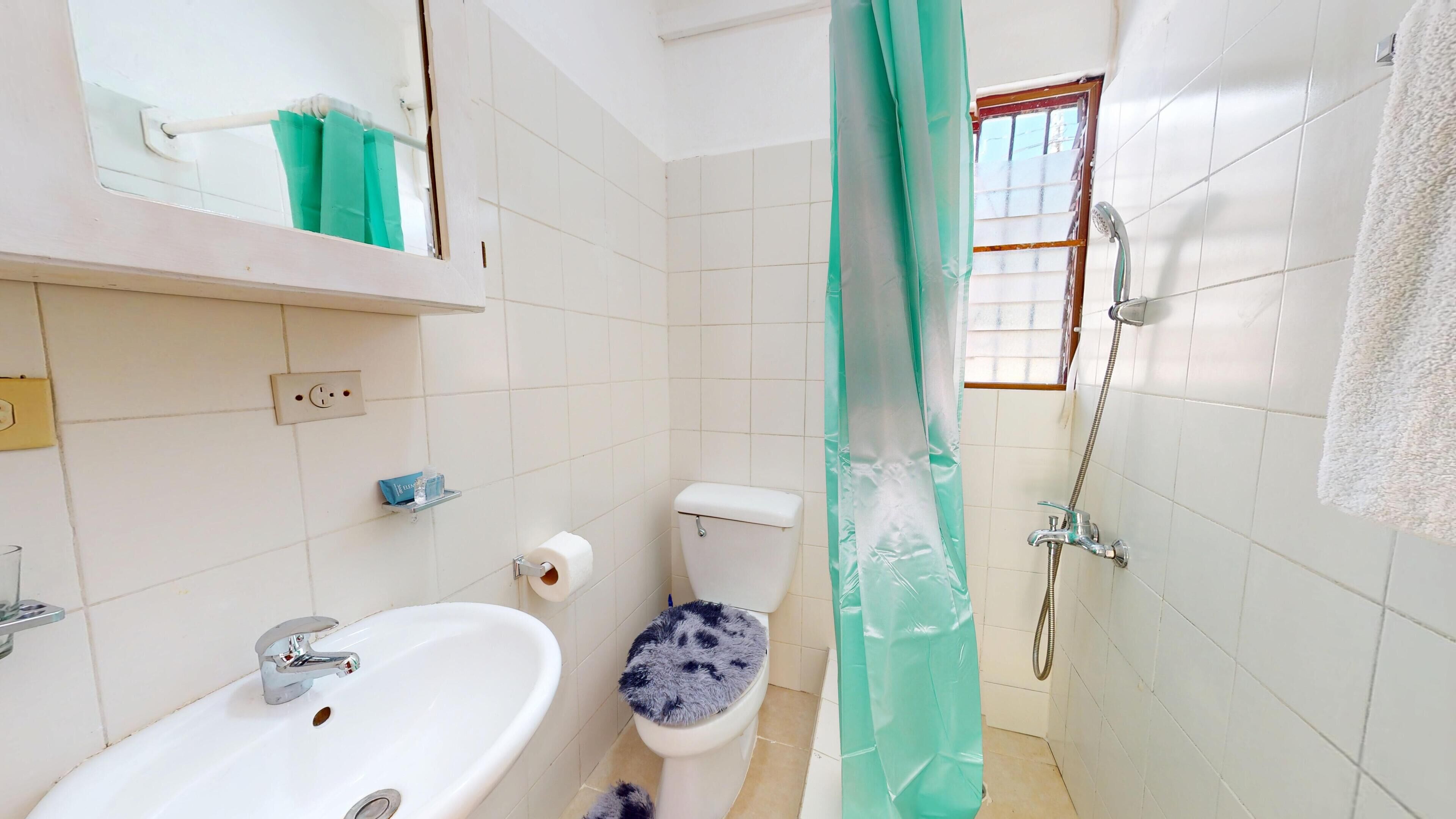 A bathroom with a white sink, toilet, and shower. There is a green shower curtain, a mirror above the sink, and a window with blinds.