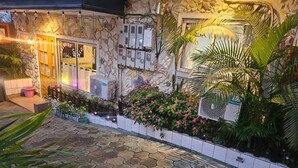 A stone building with a tiled patio, a small garden with pink flowers, and a window with a sign.