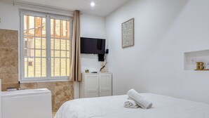 Une chambre à coucher moderne avec un lit, une armoire blanche, une fenêtre avec des rideaux, un téléviseur à écran plat et une étagère murale ornée d’une statue de Bouddha.