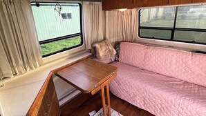 Interior compacto de um veículo recreativo, com uma mesa de madeira, um sofá acolchoado rosa e vista para uma caravana pelas janelas.