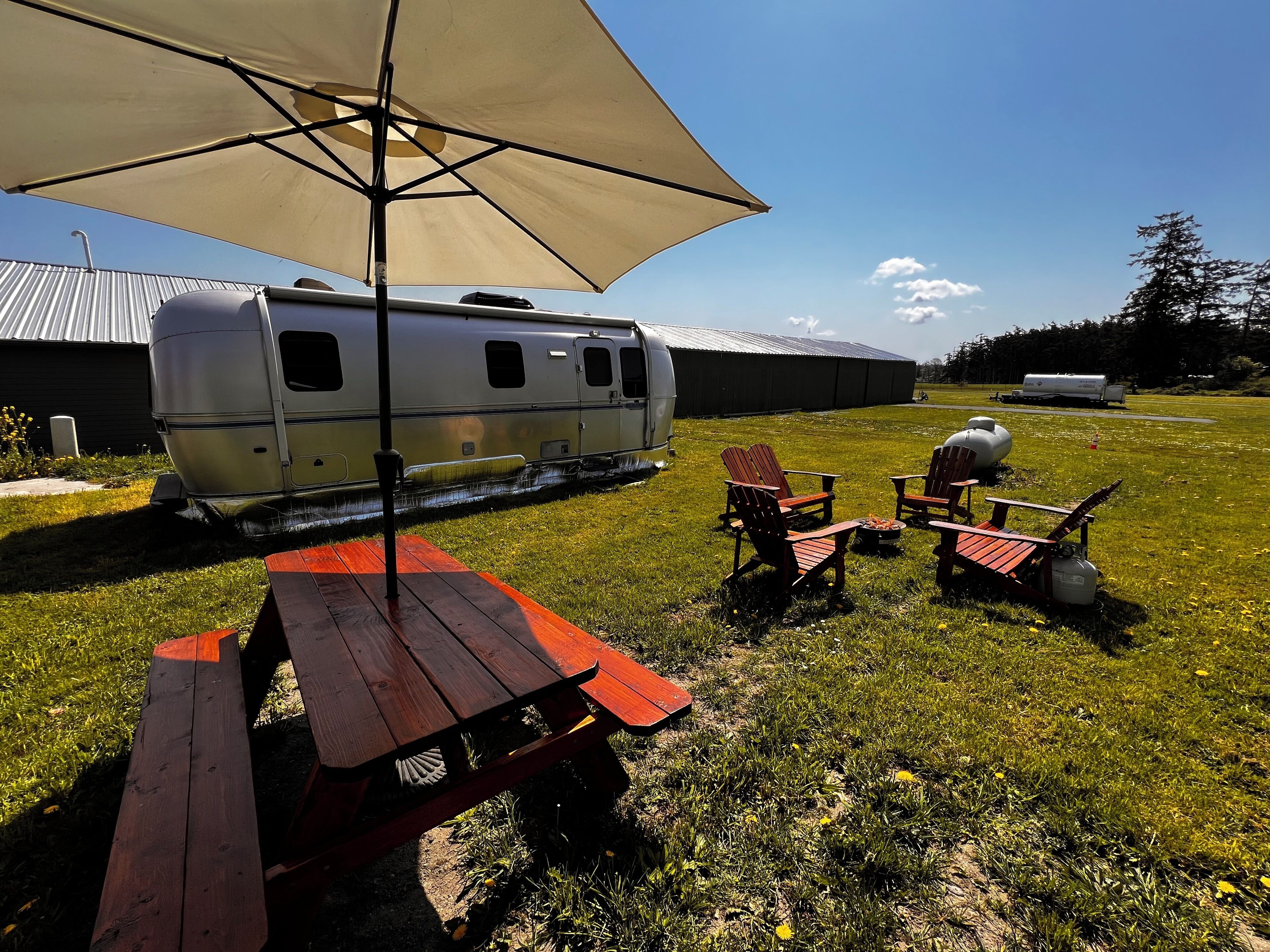 Un montaje para picnic con una mesa, sillas bajo un toldo, una caravana y una parrilla en un área con césped.