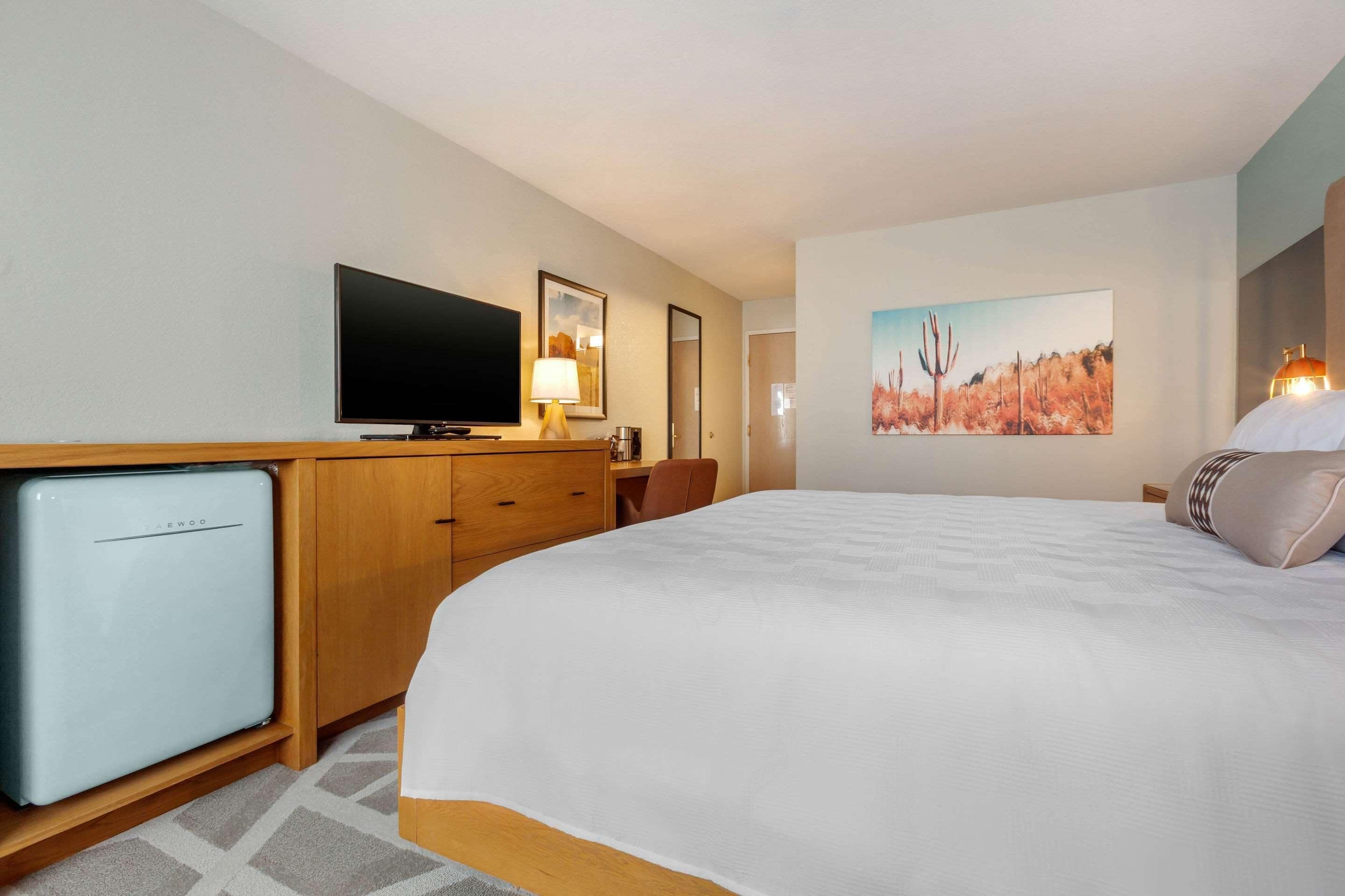 A hotel room with a bed, a television, a desk with a lamp, a refrigerator, and a painting on the wall.