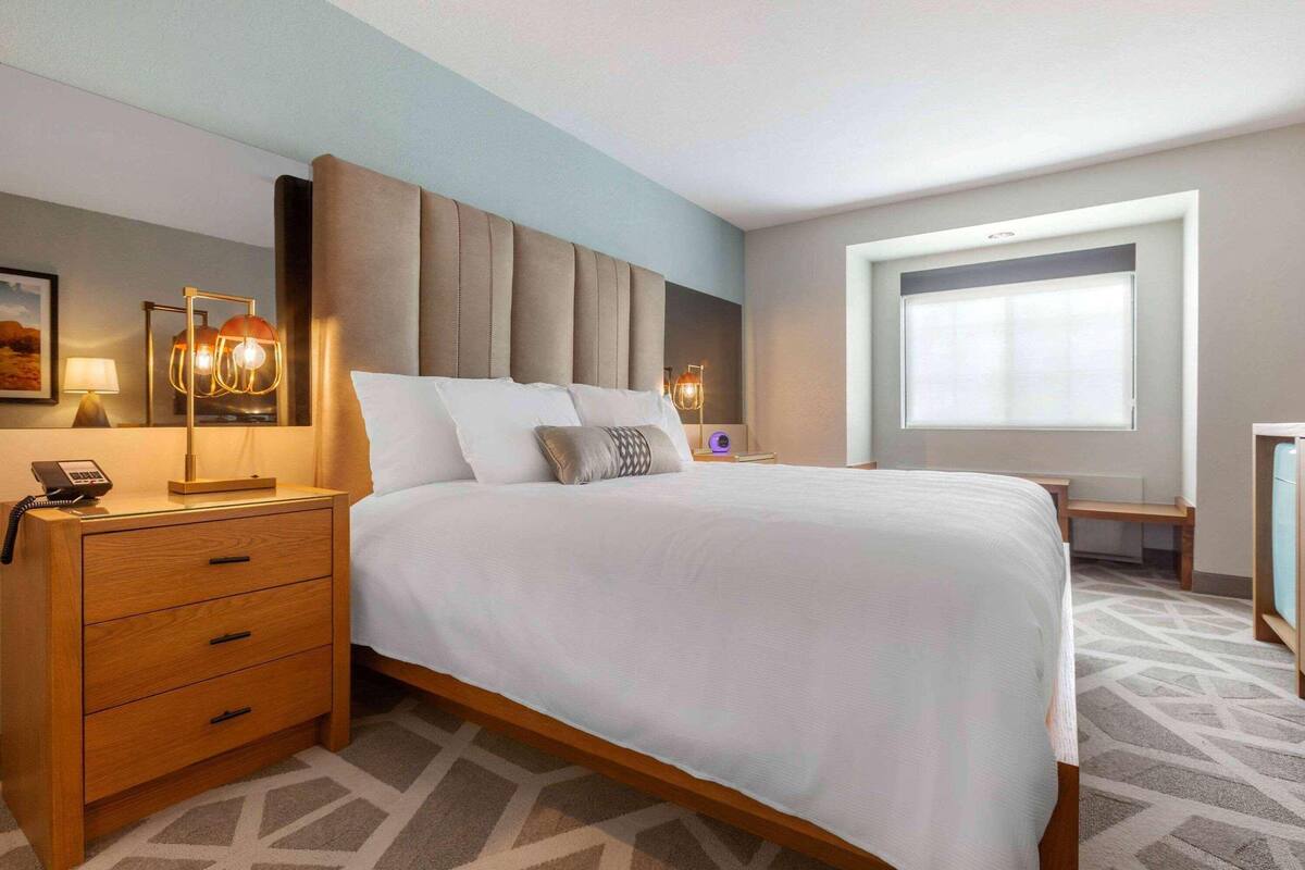 A hotel room with a large bed, a wooden nightstand with drawers, a desk, and a window with blinds.