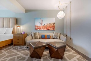 A bedroom with a bed, bedside table, lamp, sofa, ottomans, and a painting on the wall. - La Quinta Inn & Suites Tempe University / Phoenix (Tempe)