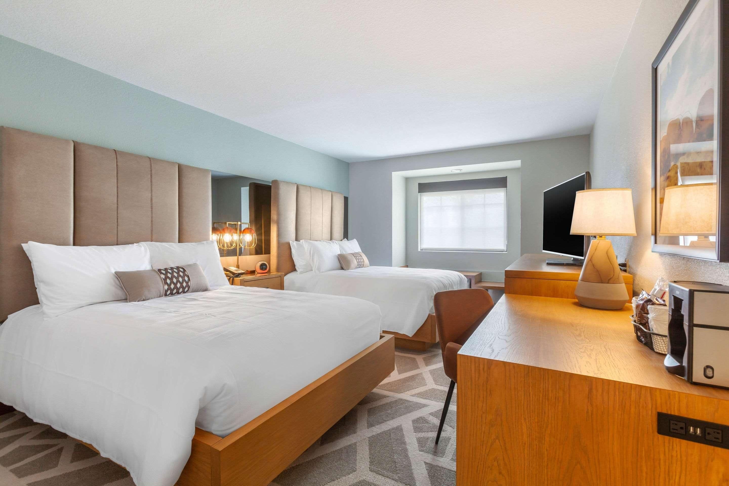 A hotel room with two beds, a desk, a chair, a TV, and a window.
