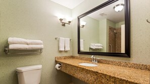 A bathroom with a granite countertop sink, a toilet, a towel rack with folded towels, a mirror, and a shower area in the background. - Comfort Suites Seabrook Kemah (Seabrook)