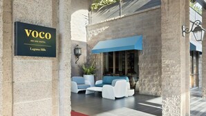 A hotel entrance with a sign reading 'Voco An IHG Hotel Laguna Hills,' outdoor seating with white and blue furniture, and a blue awning. - The Hills Hotel (Laguna Hills)