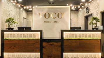 A reception desk with a sign reading 'VOCO AN IHG HOTEL' and two potted plants.