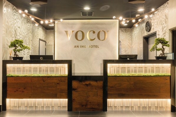 A reception desk with a sign reading 'VOCO AN IHG HOTEL' and two potted plants. - The Hills Hotel (Laguna Hills)