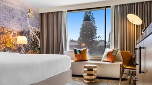 A hotel room with a bed, a sofa with colorful cushions, a small wooden table, a chair, and a large window with a view of trees and buildings. - The Hills Hotel (Laguna Hills)