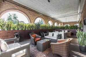Une terrasse extérieure avec des meubles en osier, une table à plateau en verre et un mur de briques orné d’arches.