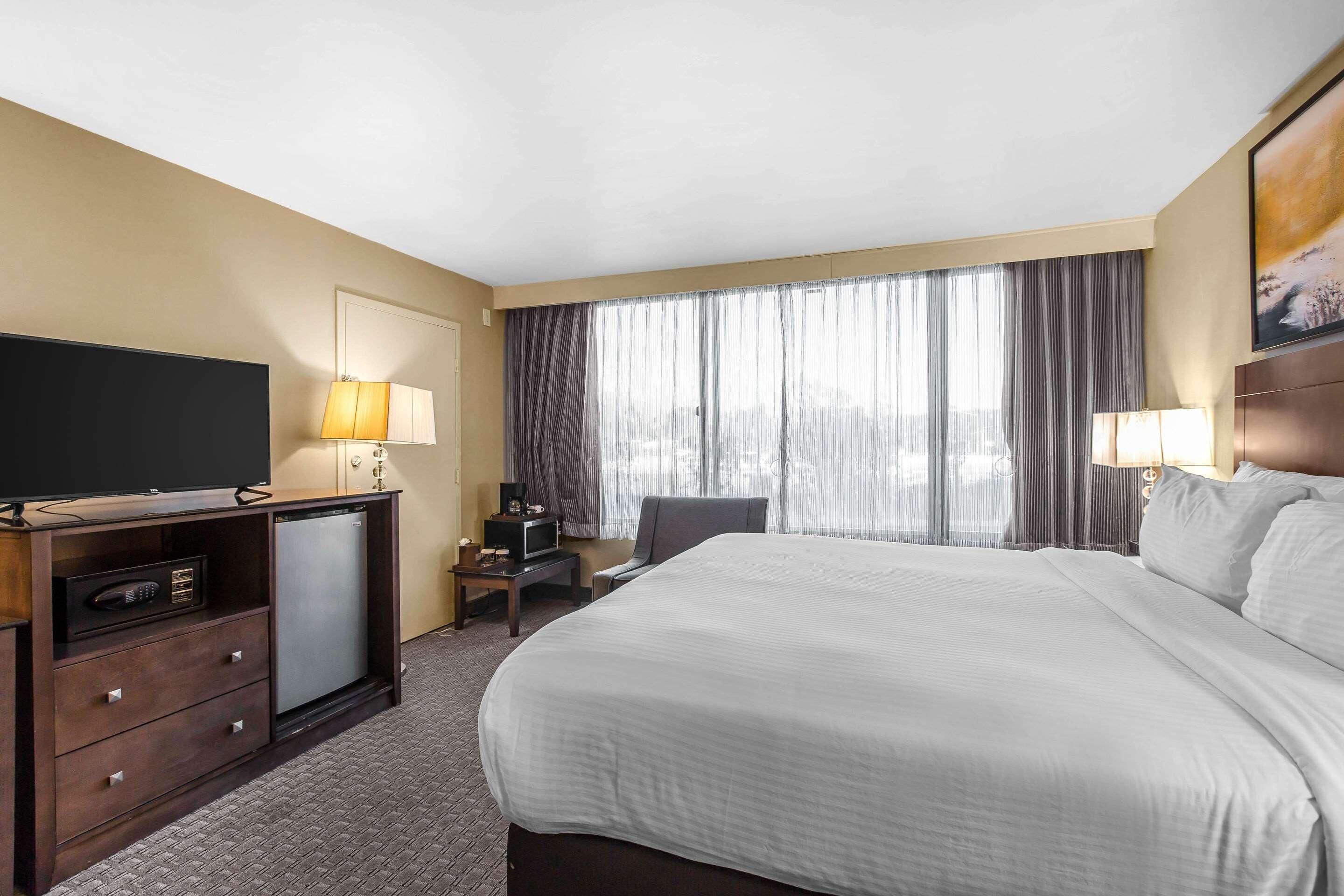 A hotel room with a large bed, a TV, a desk with a lamp, and a view of the outdoors through the window.