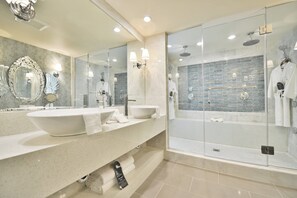 A modern bathroom with a large mirror, two sinks, a glass-enclosed shower, and a marble backsplash. - The Hills Hotel (Laguna Hills)