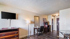 Habitación de hotel con televisión de pantalla plana, escritorio con silla, cómoda, lámpara de pie y espejo grande.