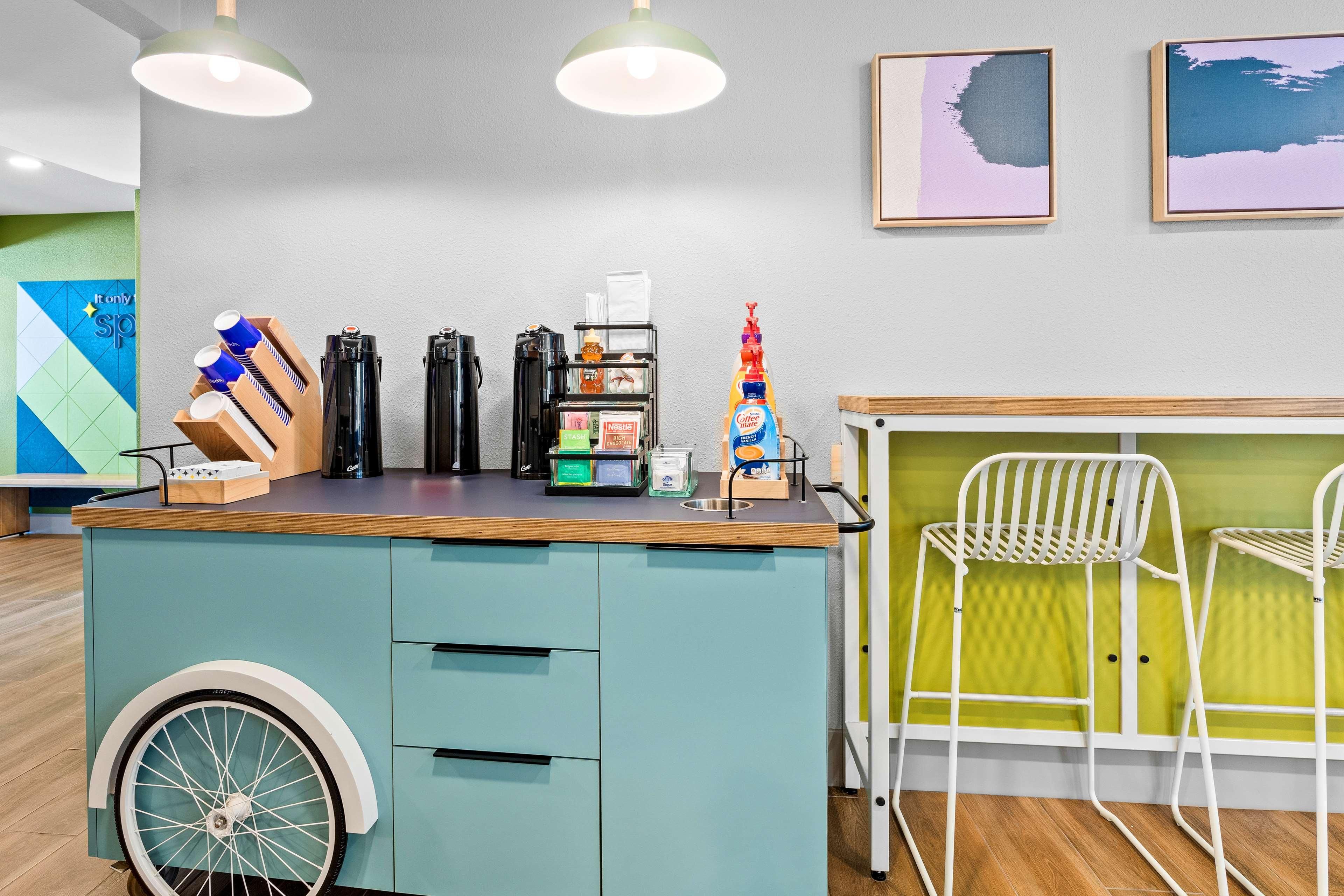 Ein Tresen mit einer Kaffeemaschine, einem Handdesinfektionsmittel und einer kleinen Pflanze. An der Wand hängen zwei gerahmte Bilder und die Wand selbst ist grün mit einem geometrischen Muster.