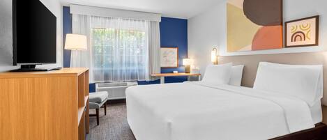 A hotel room with a bed, desk, TV, and artwork on the wall.