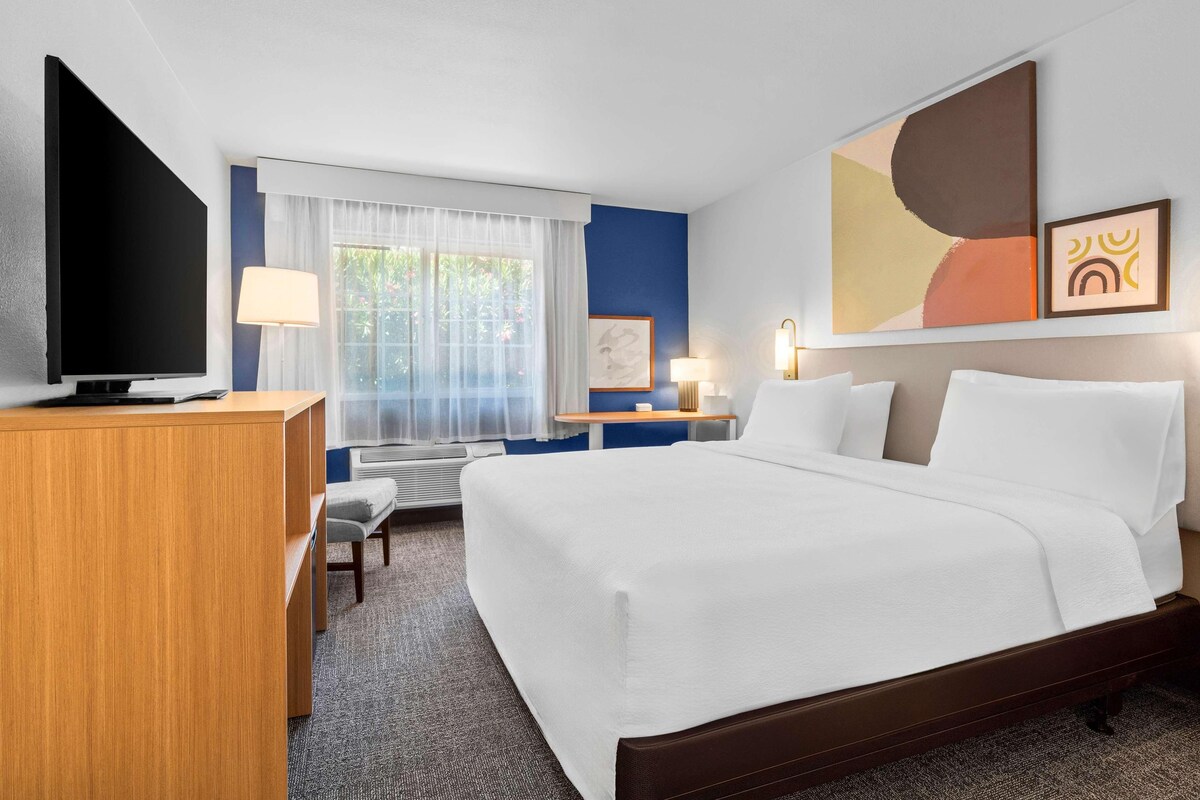 A hotel room with a bed, desk, TV, and artwork on the wall.