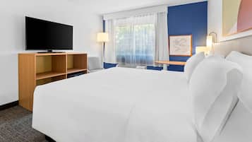 Una habitación de hotel con una cama grande, un televisor de pantalla plana sobre un soporte de madera, un escritorio con una lámpara y una ventana con cortinas translúcidas.
