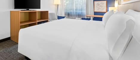 Una habitación de hotel con una cama grande, un televisor de pantalla plana sobre un soporte de madera, un escritorio con una lámpara y una ventana con cortinas translúcidas.