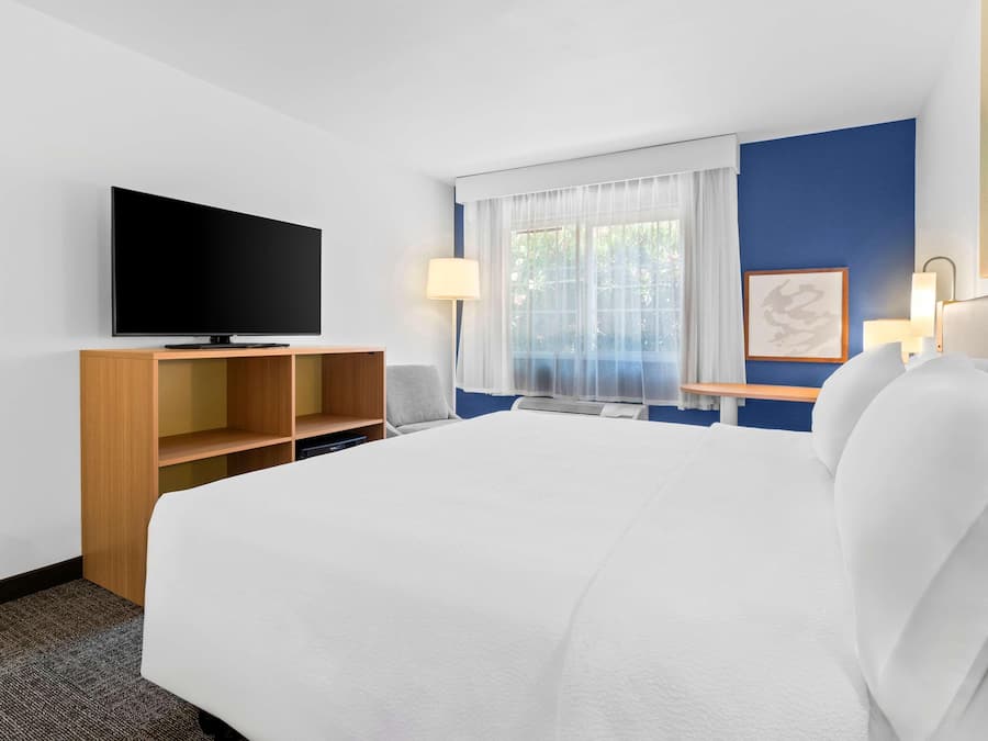 Una habitación de hotel con una cama grande, un televisor de pantalla plana sobre un soporte de madera, un escritorio con una lámpara y una ventana con cortinas translúcidas.