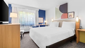 Quarto de hotel com cama, mesas de cabeceira, escrivaninha, cadeira, janela com cortinas e obras de arte nas paredes.