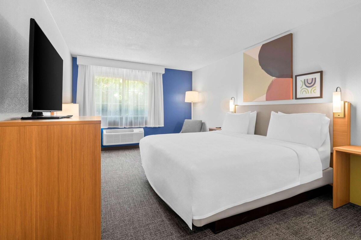 Quarto de hotel com uma cama, escrivaninha, televisão, luminária e janela com cortinas.