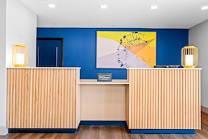 Balcão de recepção de hotel com parede azul, uma obra de arte emoldurada e duas luminárias amarelas.