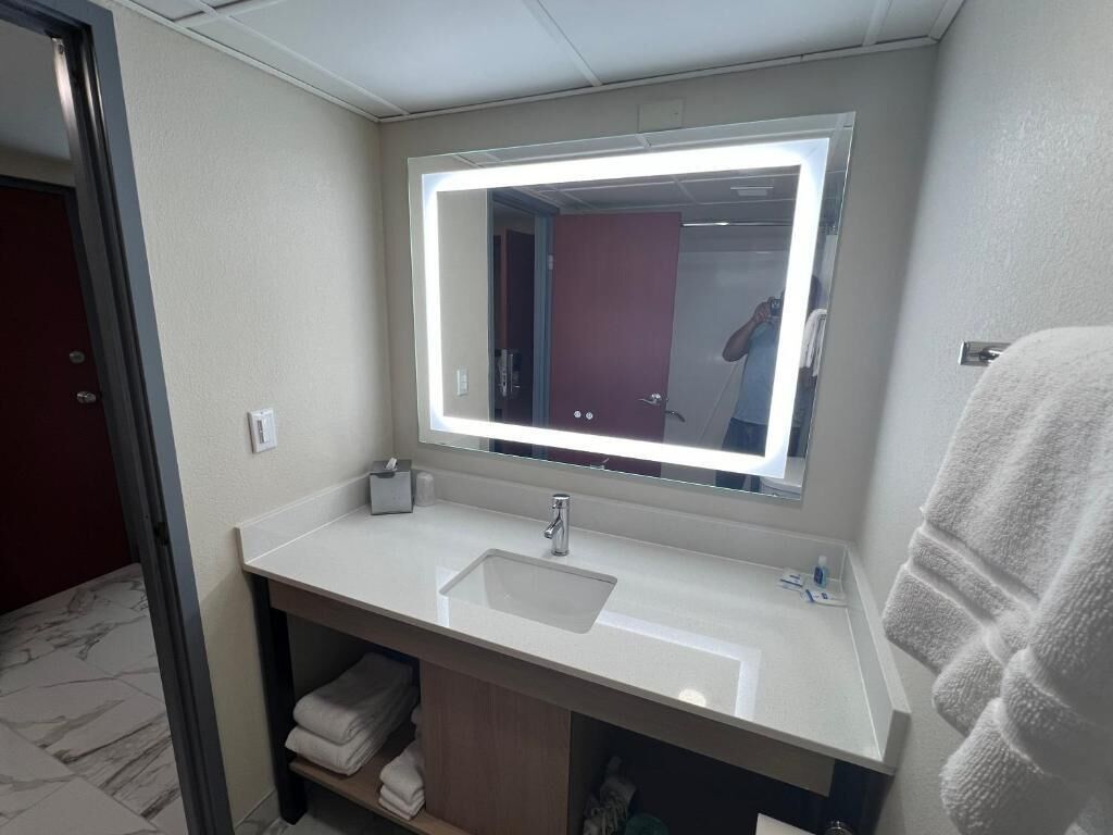 Un baño con lavamanos blanco, un espejo con luz y un perchero para toallas.