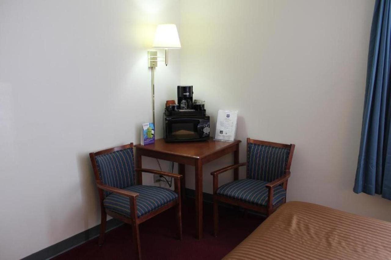 A small hotel room with two wooden chairs, a small table with a coffee maker, a lamp, and a bed.