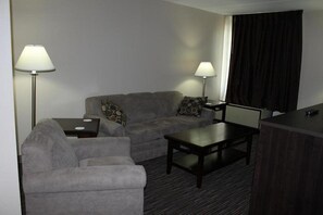 A hotel room with a grey sofa, a smaller armchair, a coffee table, two bedside tables with lamps, and a large window with dark curtains. - Rodeway Inn Little Falls NY (Little Falls)