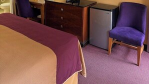 A hotel room with a bed, a desk with a television, a small refrigerator, a mirror, and a purple chair.