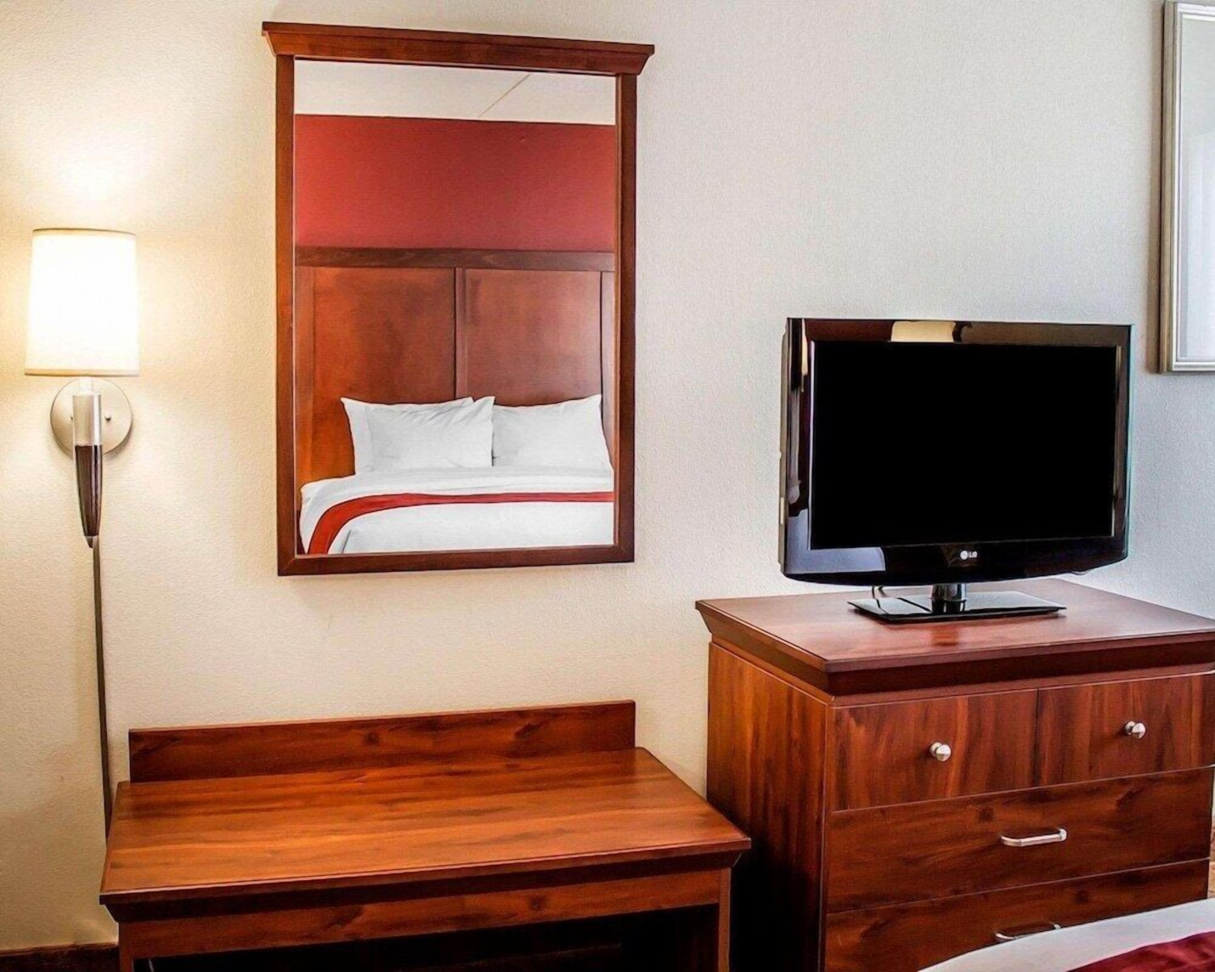 A hotel room with a wooden bed, a television on a wooden stand, a desk, and a lamp.