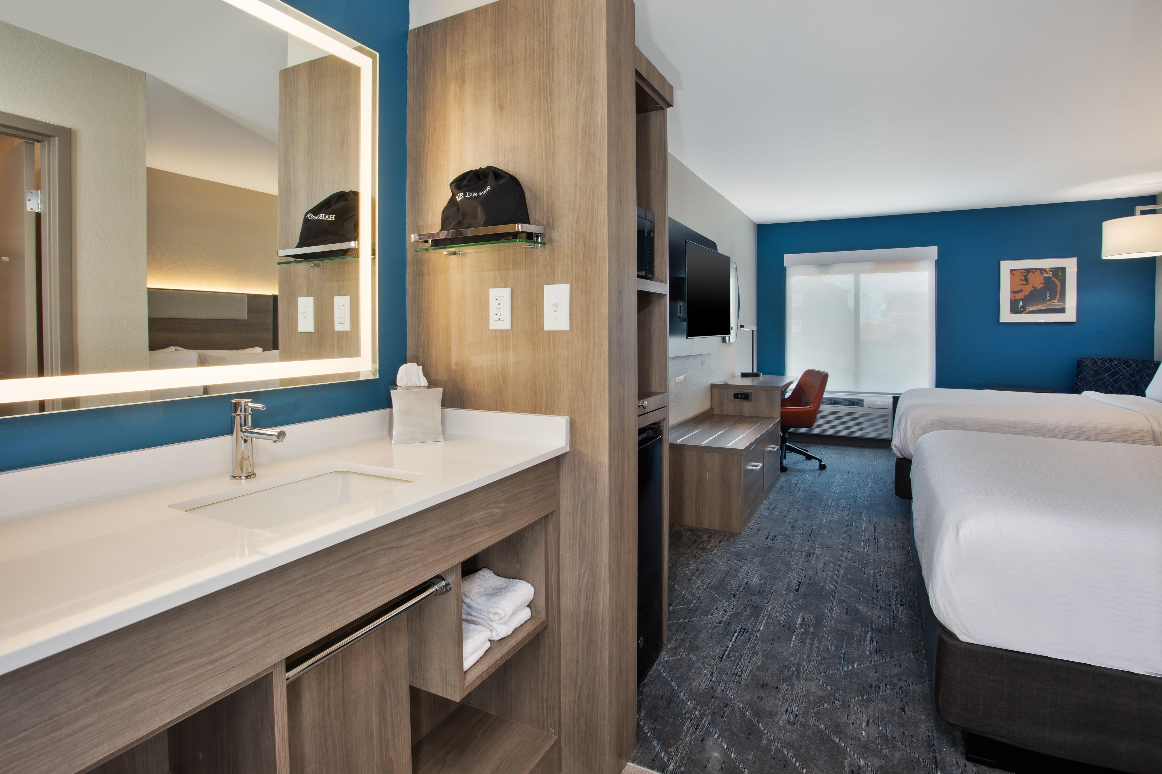 A hotel room with a large mirror, a sink, a bed, a desk with a chair, and a television.