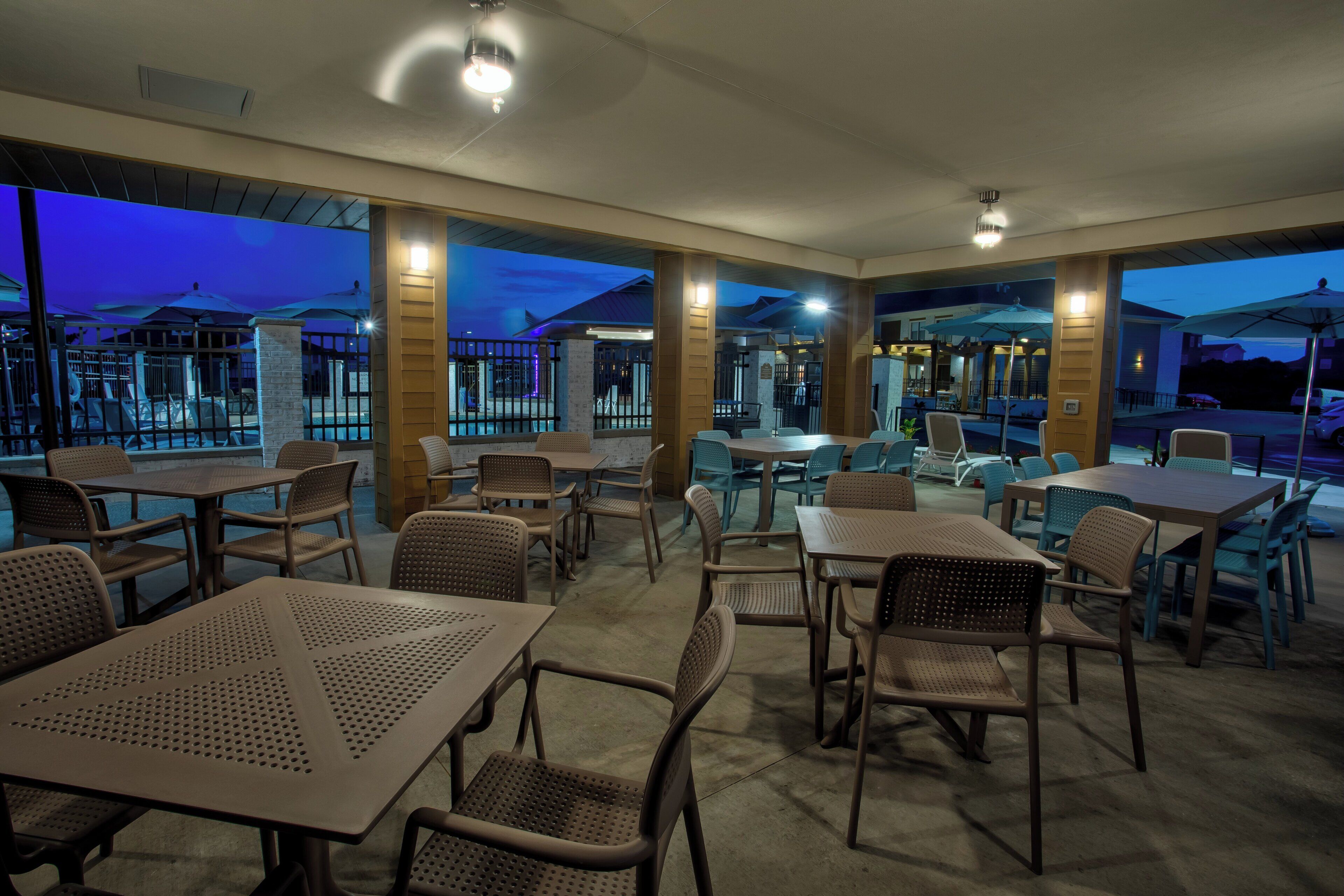 An outdoor restaurant area with tables and chairs, large umbrellas, and a view of the pool area.