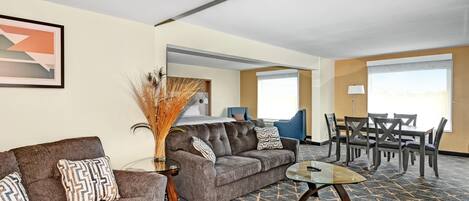 A hotel room with a sofa, armchair, coffee table, dining table, chairs, and a large window.