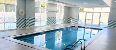 Una piscina interior con agua azul cristalina, una escalera de piscina y vistas al exterior a través de grandes ventanales.