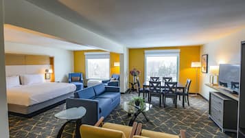A hotel room with a bed, sofa, dining table, chairs, TV, and a small table with a plant.
