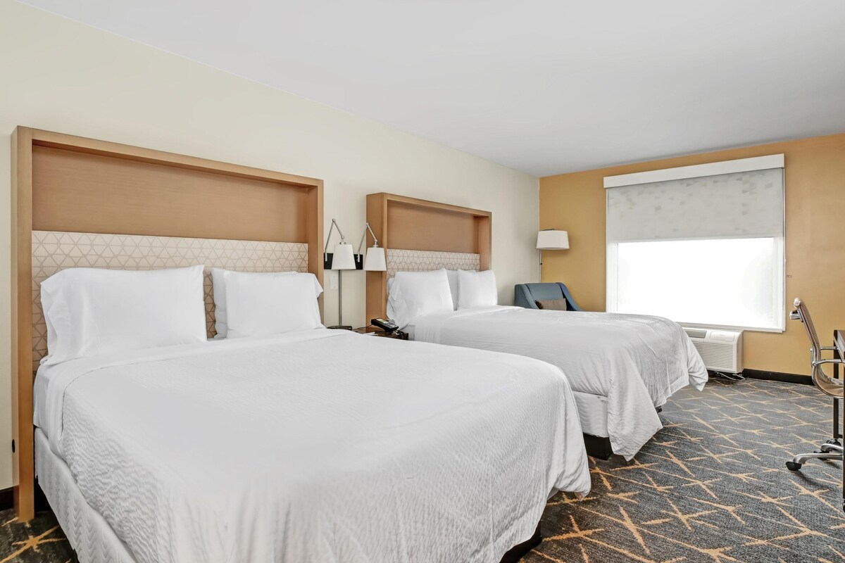 A hotel room with two beds, a desk, a chair, a lamp, and a window with a view.