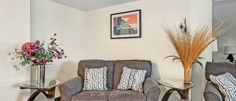 A modern living room with a grey sofa, two patterned pillows, a glass vase with flowers, and a framed abstract painting on the wall.