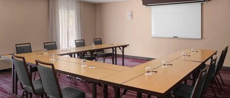 A conference room with a long table, chairs, a projector screen, and a window with curtains.