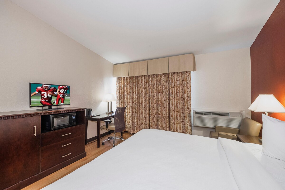 A hotel room with a bed, a desk with a chair, a television, a lamp, and an air conditioning unit.