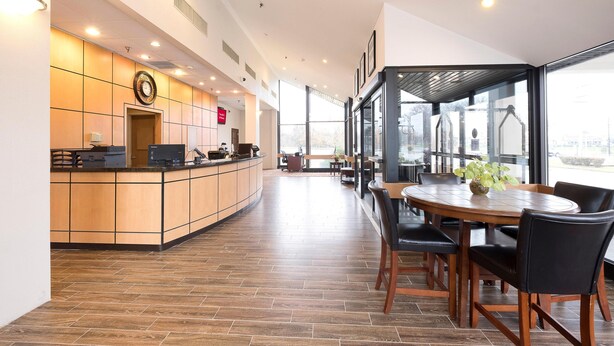 Un hall d’hôtel moderne comprenant un comptoir d’accueil, un espace repas avec une table ronde et des chaises, ainsi que de grandes fenêtres offrant une vue sur l’extérieur.
