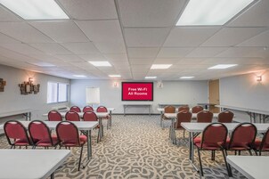 Un salón de conferencias con mesas y sillas, una pantalla de proyector que muestra 'Wi-Fi gratuito en todas las habitaciones' y una alfombra estampada.