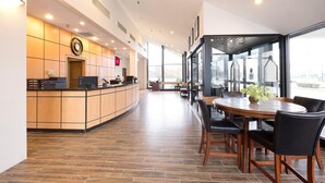 A modern hotel lobby with a reception desk, a dining area with a round table and chairs, and large windows providing a view of the outdoors.