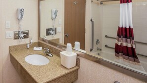 A bathroom with a granite countertop sink, a mirror, a hairdryer, a shower with a grab bar, and a towel rack.