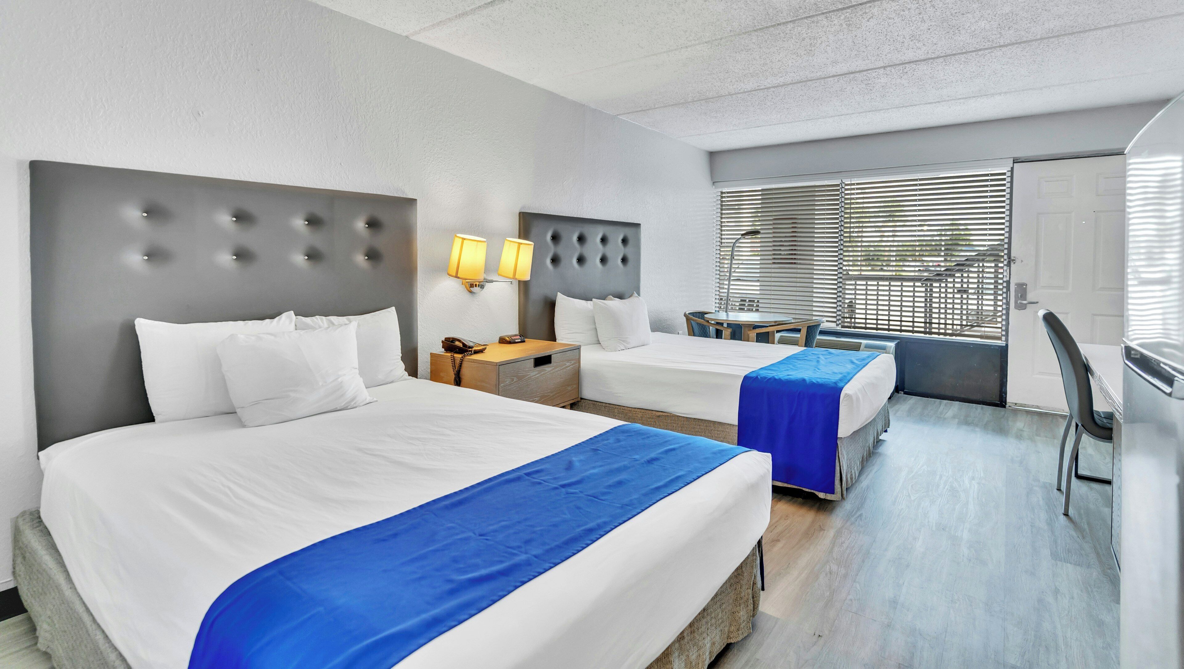 A hotel room with two beds, a desk, a chair, and a window with blinds.
