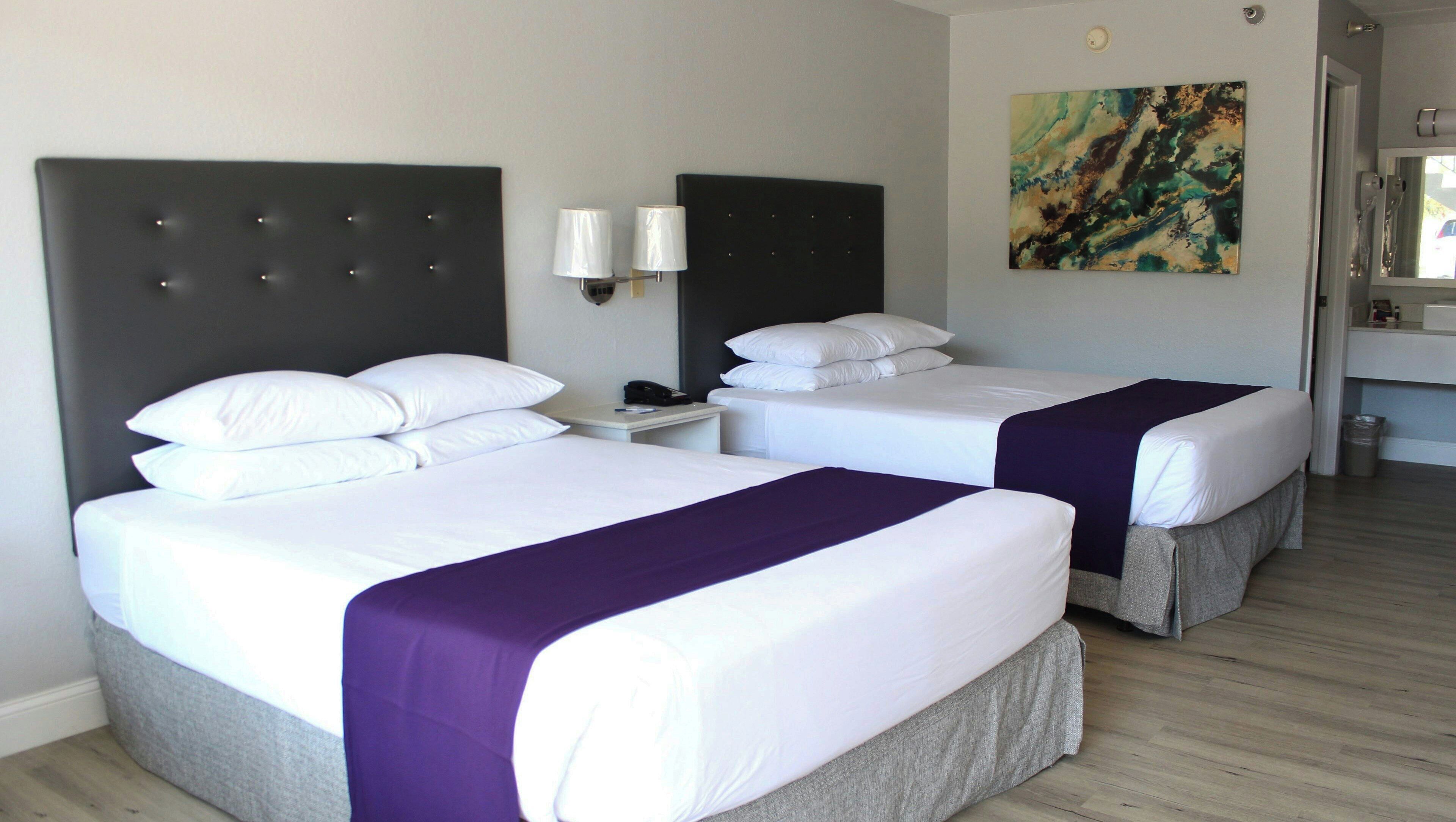 A hotel room with two beds, a nightstand, a lamp, a painting, and a bathroom visible through an open door.