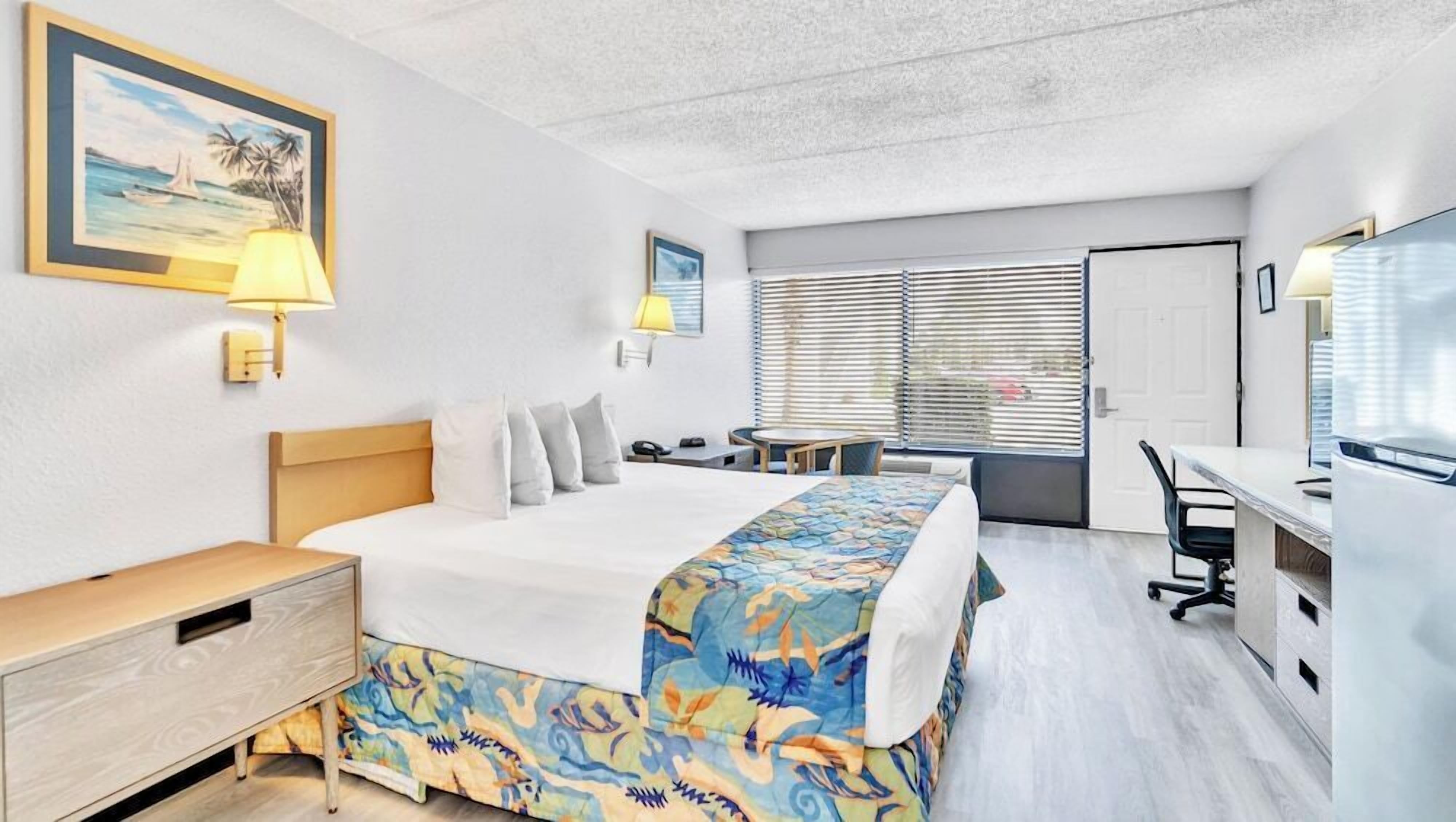 A hotel room with a bed, bedside table, lamp, desk, chair, and a window with blinds.