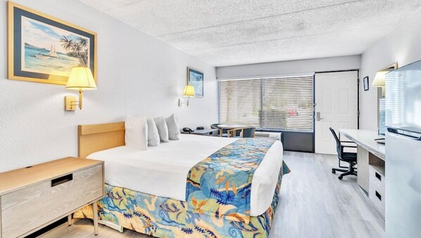 A hotel room with a bed, bedside table, lamp, desk, chair, and a window with blinds. - Home 1 Suites Extended Stay - Kissimmee (Kissimmee)